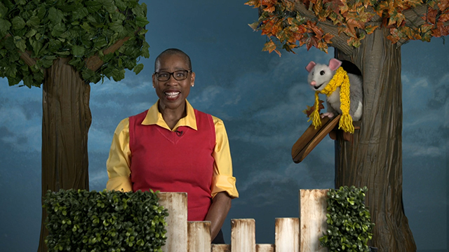 Black woman with short hair smiles, stands next to puppet possum with yellow scarf in tree