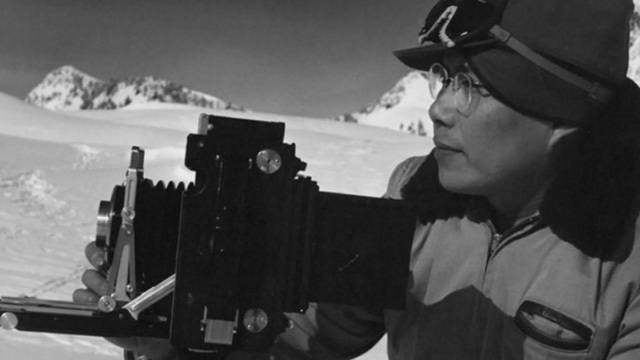 Black and white image of man's shoulders and face looking toward large camera, snowy mountain background