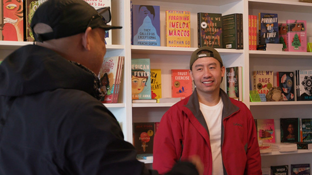 Cambodian American man in red jacket and backwards ball cap smiles at man with back to camera, shelves of books behind