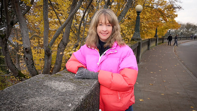 Nancy Guppy smiles at camera in pink and orange puffer jacket, leans on cement wall with street and autumn leaves in background