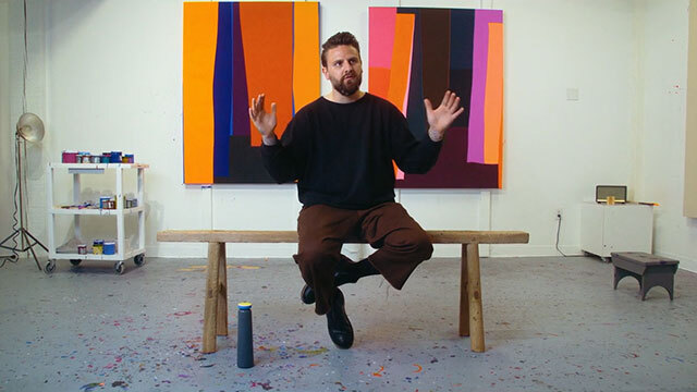 Brian Sanchez sits on bench with hands raised in front of two multicolored abstract paintings.