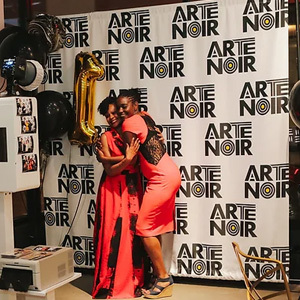 Two black women dressed to the nines and getting their picture taken with a party backdrop and gold balloons