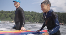 An adult and an Indigenous youth smile as they walk a surf board out into the ocean