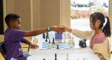 Two children, one Black and one Asian, shake hands from across the table over a chess game. 