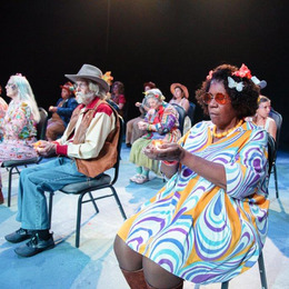 A group of diverse performers sit on chairs on a stage holding their hands out in front of them.