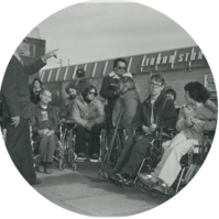A black and white photo of a group of people in wheel chairs listening to a person in a hard hat speak.