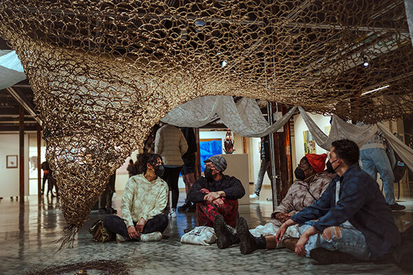 Four people wearing facemasks sit crosslegged under a large woven lattice that hangs low above them.