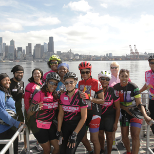 A group of pedal bikers pose together on the front of the water taxi with the Seattle city skyline behind them. 