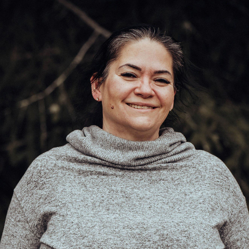 A Native woman smiling, wearing a gray sweater with trees in the background.
