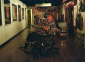 A Black man in a wheelchair has one arm raised, reciting a monologue from a stage play.