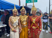 Three people wearing traditional Cambodian attire stand together at a street fair. 