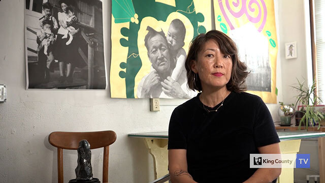 Erin Shigaki sits in front of art featuring various people