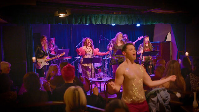 Theater with live band and people singing onstage, performer in sparkly tank top stands amongst audience members