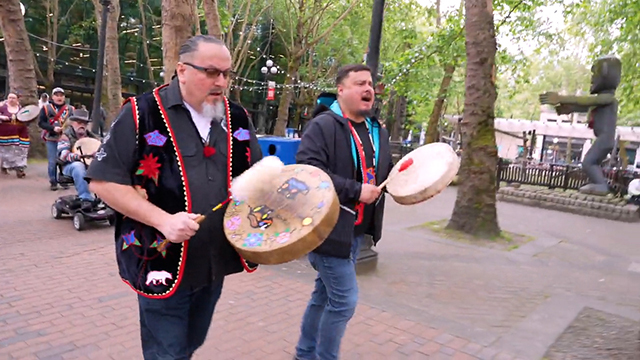 Two people in foreground walk while hitting handheld drums, others follow behind