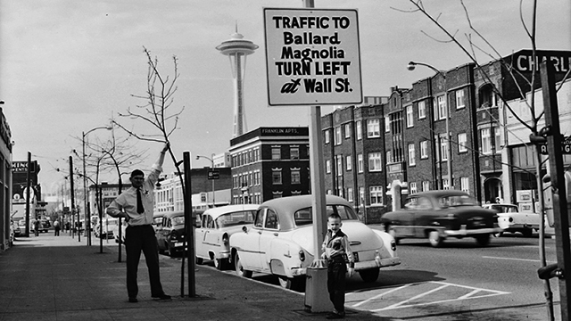 Black and white image of man holding sapling in one hand, street and Space Needle in background