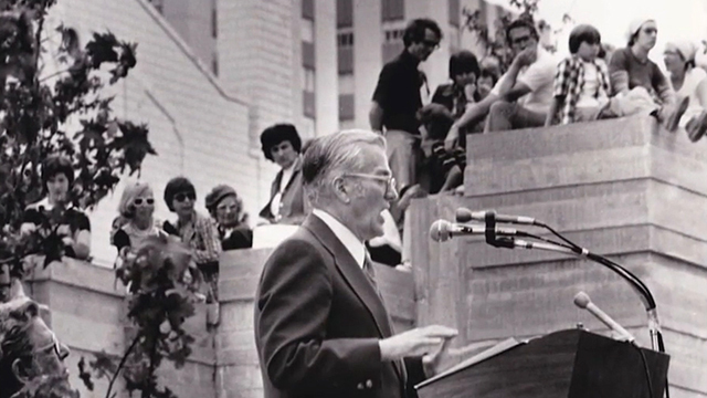 Black and white image of Jim Ellis speaking into mic at podium, in background, people watch from above