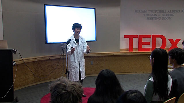 Kid in white lab coat stands in front of room with red Tedx sign behind