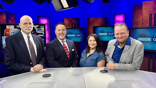 Four people stand side by side behind white desk with blue background in television studio