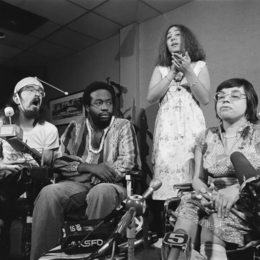 A black and white photo of three people in wheelchairs speaking into news microphones 