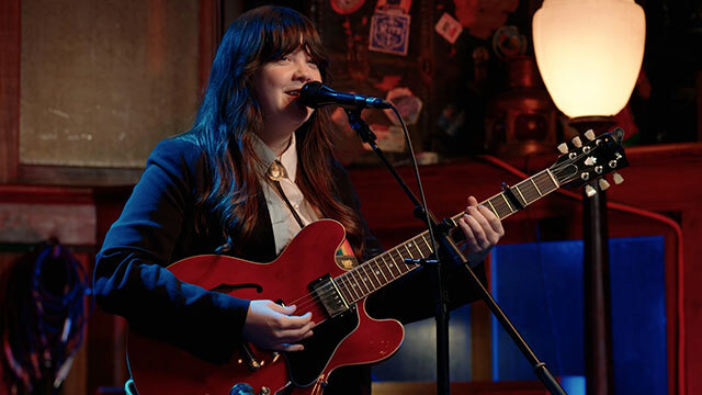 K Van Petten stands onstage behind microphone holding guitar