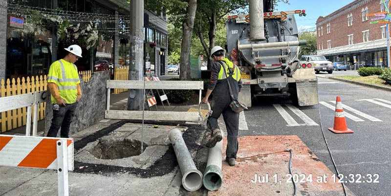 Two construction workers in yellow reflective vests and white helmets stand above a new pothole for the custom signal pole.  