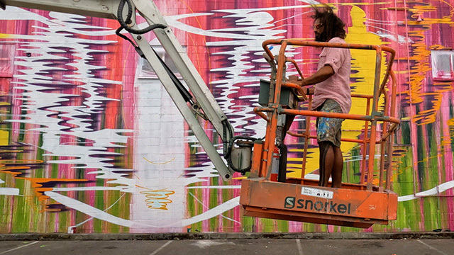 Person stands in cherry picker in front of pink and green mural