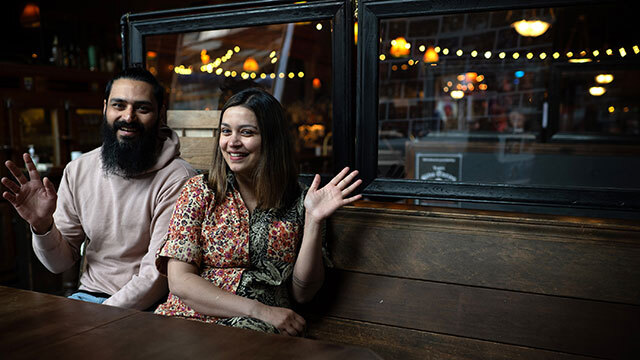 Man and woman sit on wooden bench, smiling and waving at camera 