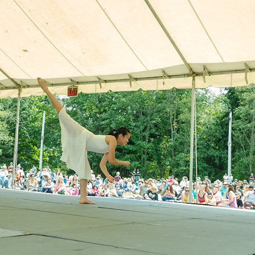 A dancer wearing a flowy white dress pictured leaning forward with one leg kicked high in the air.