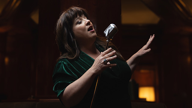 Woman in green velvet leans back, singing into retro-style microphone