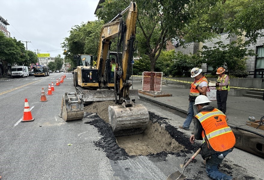Photo depicts construction workers on the road near an excavator. 