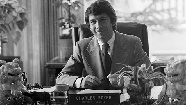 Black and white photo of Charles Royer smiling at desk, holding pen