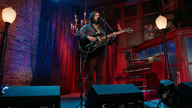 Man holding guitar onstage sings into mic