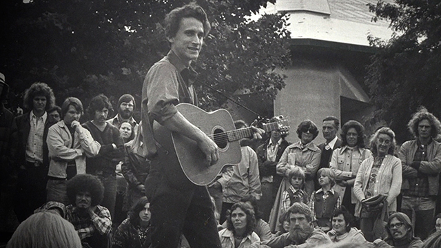 Black and white image of man playing guitar as people around watch