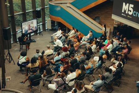 A seated crowd watching a moderator and presenter on a screen