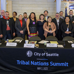 Eighteen people stand behind a table with a tablecloth with text that reads: "City of Seattle Tribal Nations Summit, May 2, 2023."