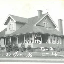 A vintage black and white photo of a house on a corner with a wrap around deck and a sign with letters "YWCA"