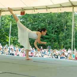 A dancer wearing a flowy white dress pictured leaning forward with one leg kicked high in the air.