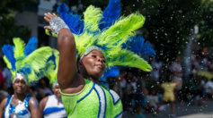 A performer wearing a green and blue leotard and feather headband dances in a street parade.