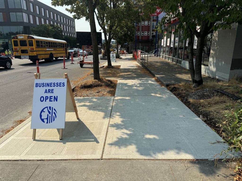 A businesses are open sign set alongside the newly installed sidewalk pavement on 15th Ave NW.