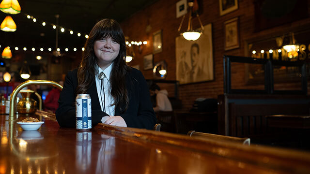 Person sits smiling at bar in white shirt, black jacket, bolo tie. Can on bar in front of them.