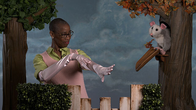 Black woman with glasses wears long pink satin-textured gloves, shows to possum puppet in tree