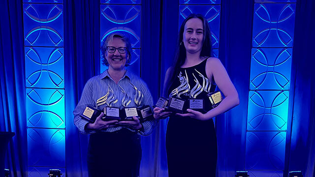 Two women stand in front of blue background holding trophies in both arms