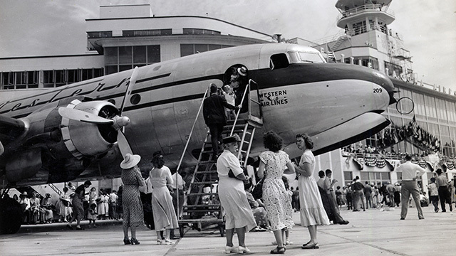 Black and white image of people getting on small plane, others milling about on tarmac with airport terminal behind