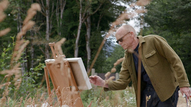 Man stands in field painting at easel, trees in background