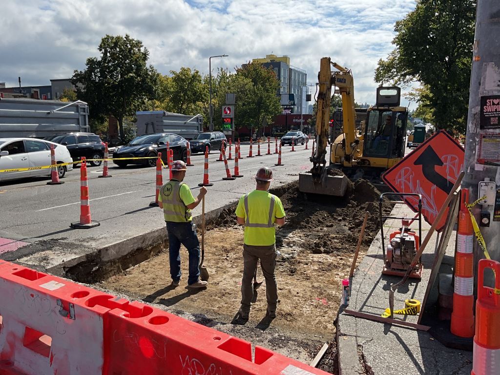 Two construction crew members oversee a large excavator as it digs up dirt to access utilities under the street.