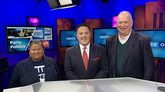 Three people stand smiling in blue television studio