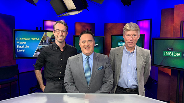 Three people stand behind white desk in blue television studio