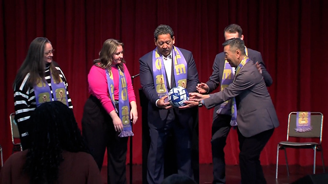 Five people including Mayor Harrell stand onstage in purple scarves, mayor and two others hold soccer ball