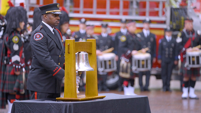 Firefighter stands at large silver bell, others in background