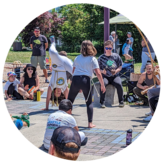 A group of people sit in a circle in a park with two people in the center doing dance and acrobatic moves